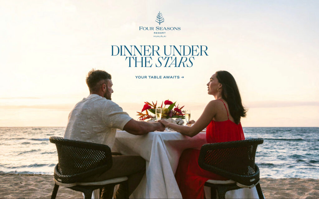 A couple enjoys a romantic Big Island Hawaii Dinner Under The Stars at the Four Seasons Hualalai, holding hands at a beachside table as sunset paints the sky and ocean in a tranquil, picturesque scene.