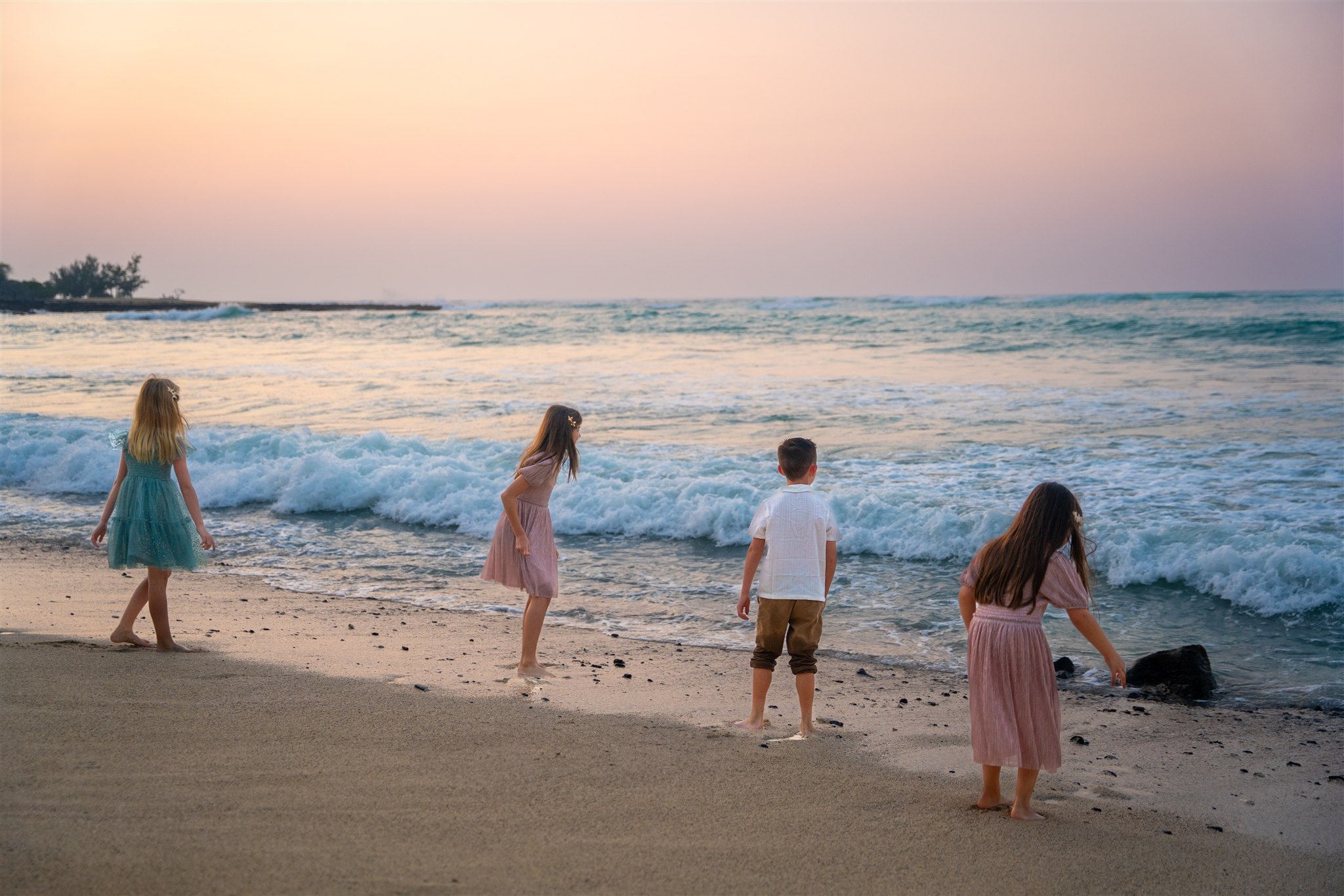 Four children, three girls in dresses and one boy in a t-shirt and shorts, play barefoot on a sandy shore at sunset&mdash;capturing the joy of hawaii family beach photos with waves rolling in under a glowing sky.