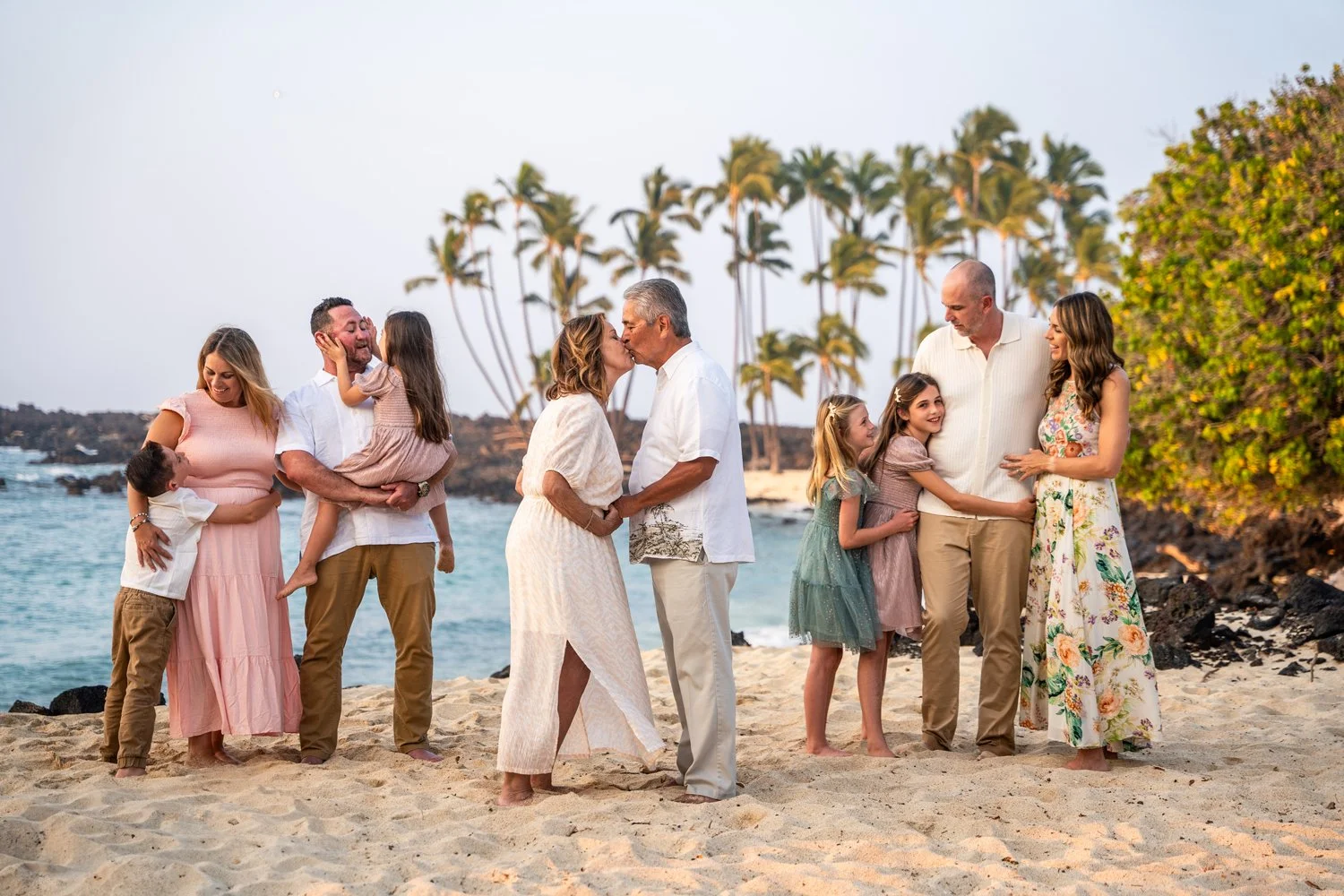 Three couples with children stand together on a sandy beach with palm trees and the ocean in the background, posing and embracing each other.