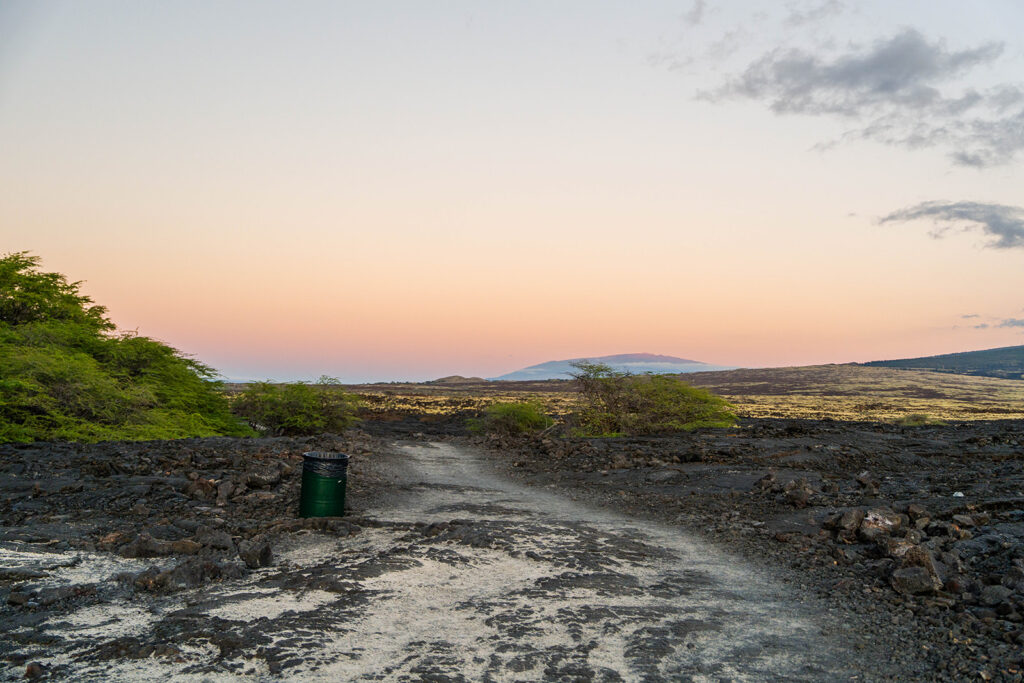 A dirt path runs through dark volcanic rocks and sparse green bushes under a pastel sunset sky, with a green trash can on the side and distant mountains in the background.