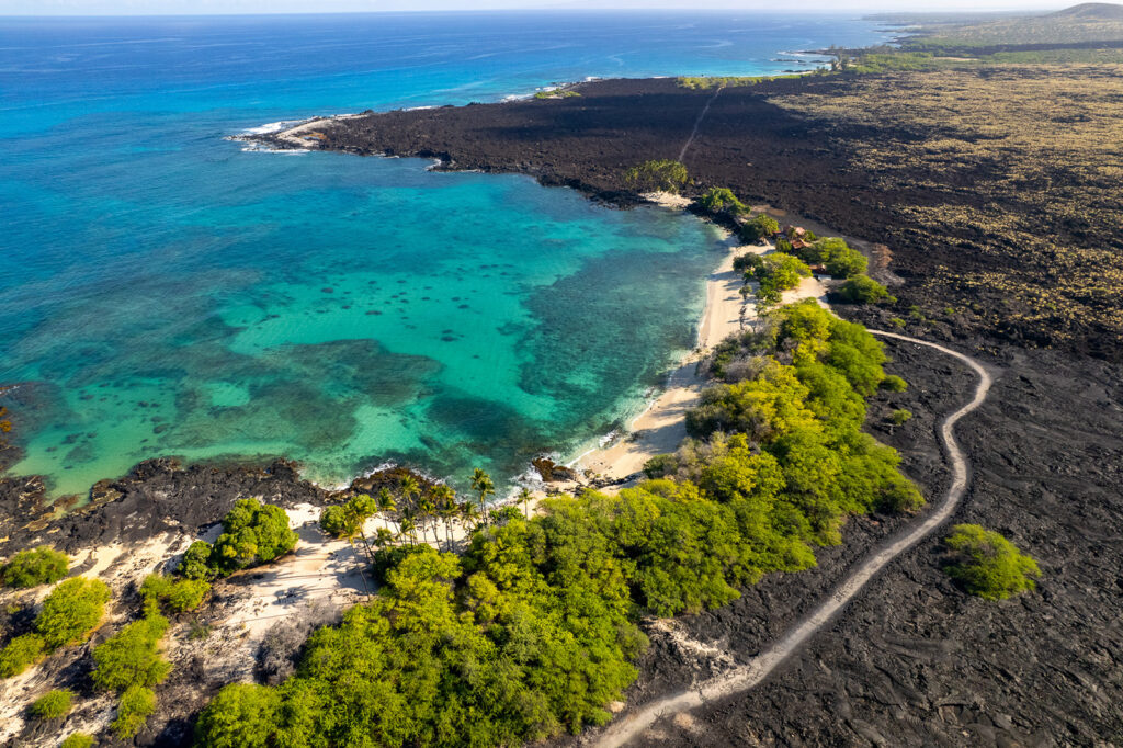 Aerial view of a coastal area with turquoise water, a sandy beach, lush green trees, and a winding path surrounded by dark volcanic rock and sparse vegetation.