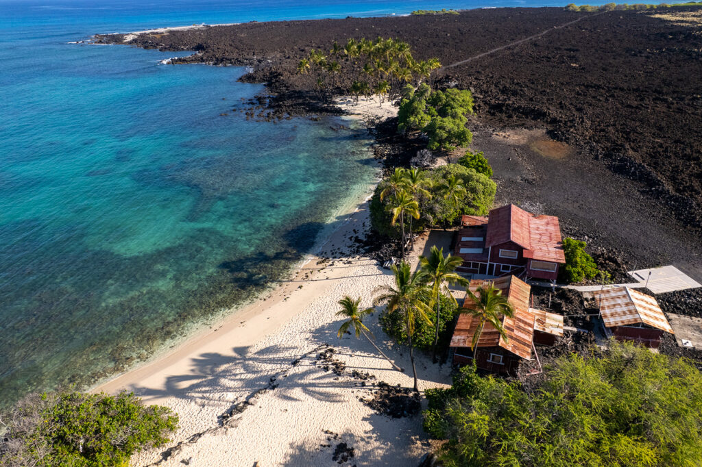 Aerial view of rustic red beachfront houses on white sand, surrounded by palm trees, black volcanic rocks, and clear turquoise ocean water.