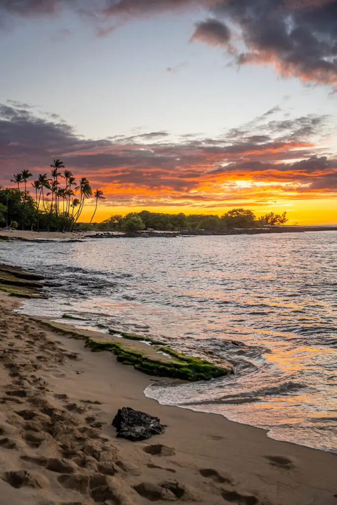 A sandy beach with footprints leads to gentle ocean waves at sunset—perfect for family photos. The sky glows orange and purple, while palm trees line the distant shore under dramatic clouds, capturing memories with a Big Island family photographer.