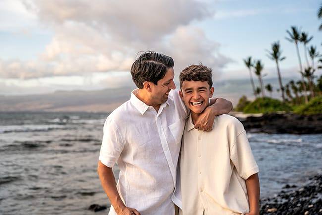 Father and teenage son laughing together during a Hawaii family photo session by the ocean.
