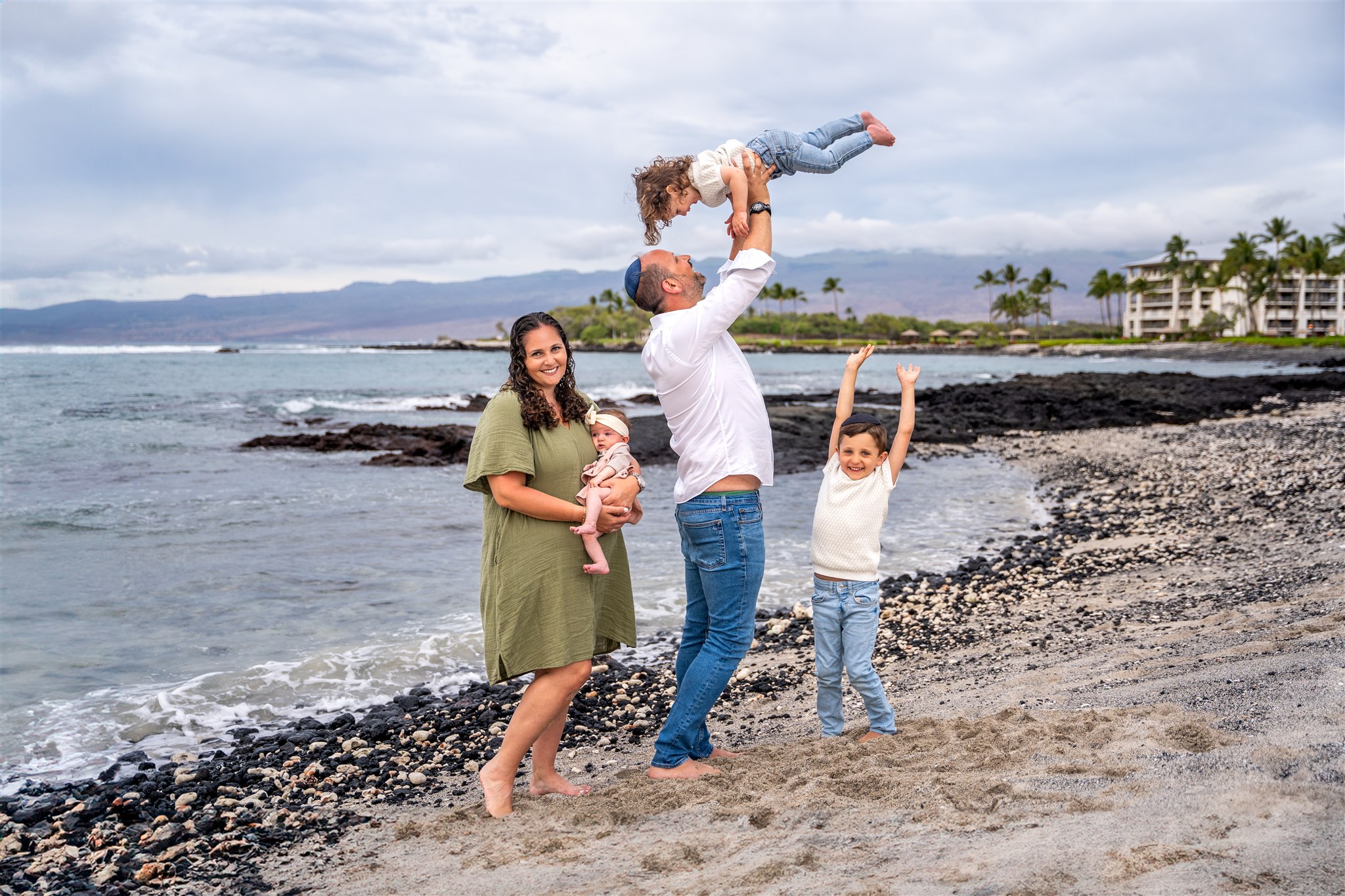 Dad holds his kid in the air with his family surrounding him in Hawaii on the beach.