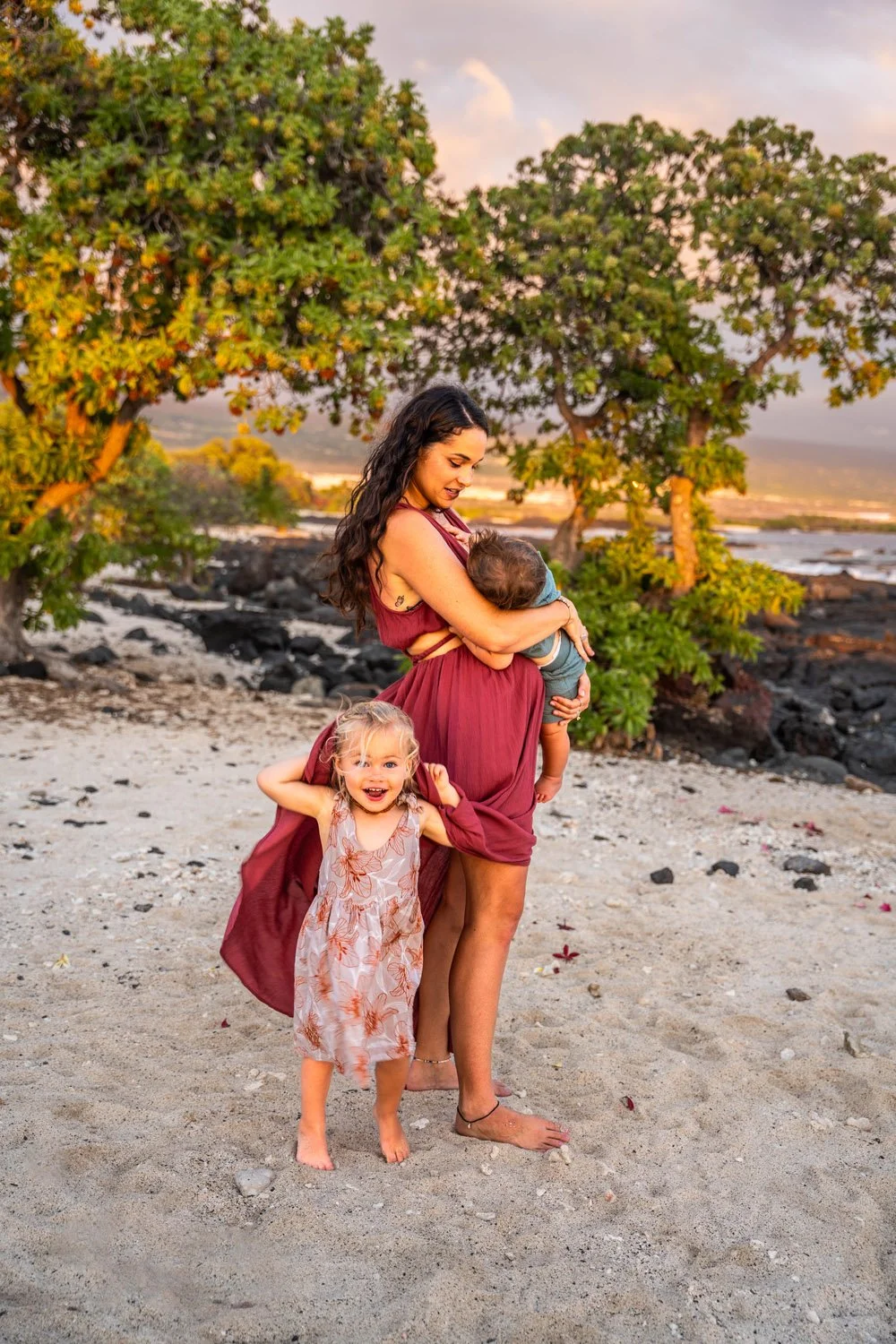 mother holding baby boy while her toddler girl hides under her dress at a white sandy beach on the big island of hawaii