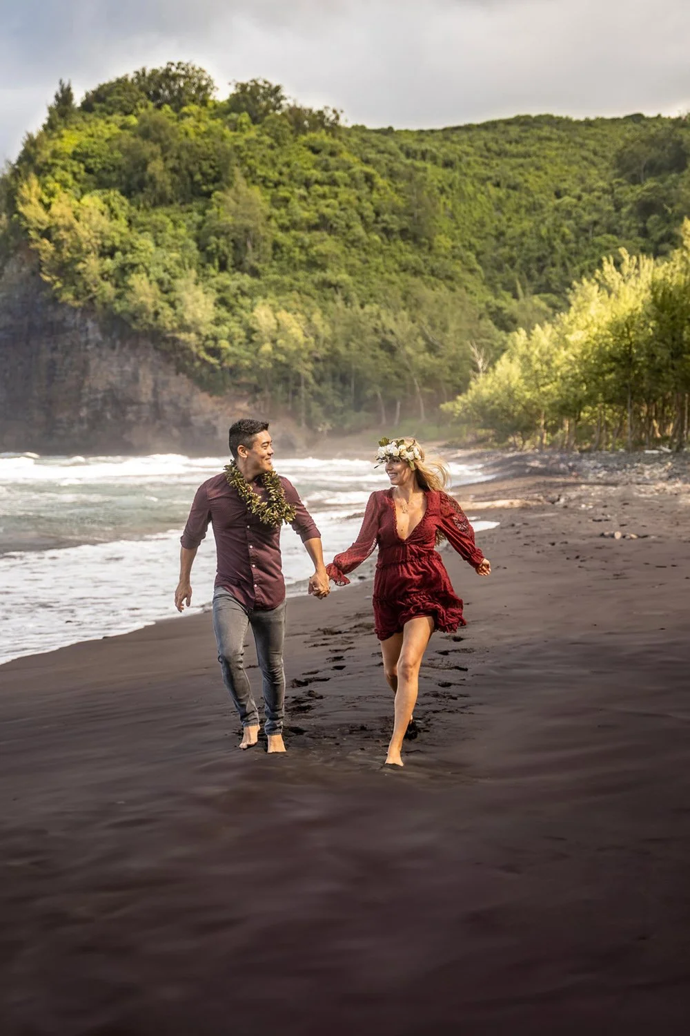 couple running holding hands on a black sand beach with a valley backdrop for a couples portrait session on the Big Island of Hawaii