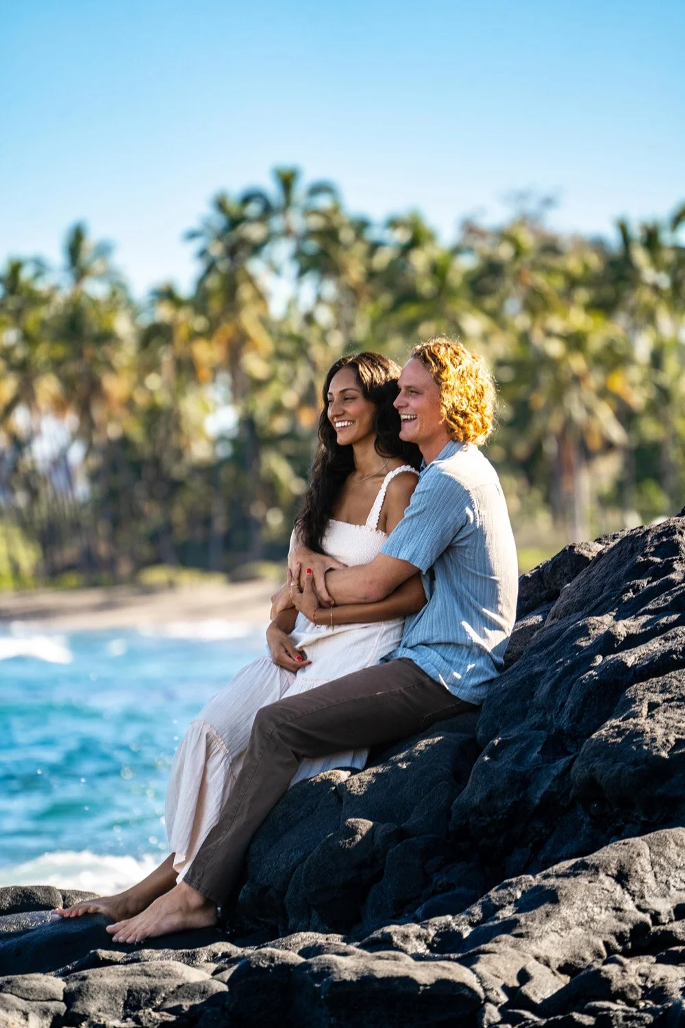 couple holding each other sitting in boyfriend's lap on lava rock with palm trees in the background for a couples portrait session on the Big Island of Hawaii