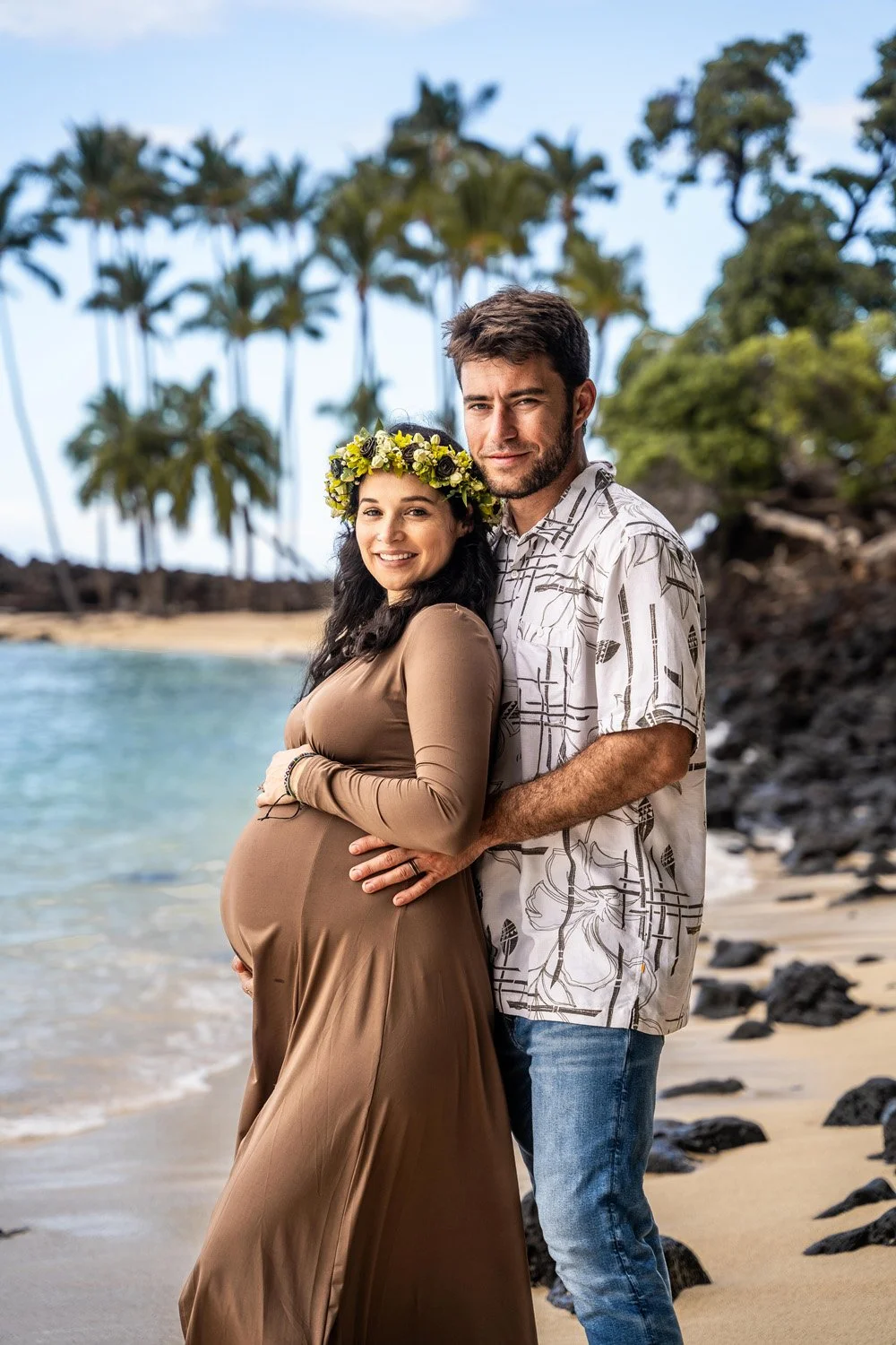 couple holding belly for a babymoon maternity session on a white sandy beach with palm trees on the Big Island of Hawaii