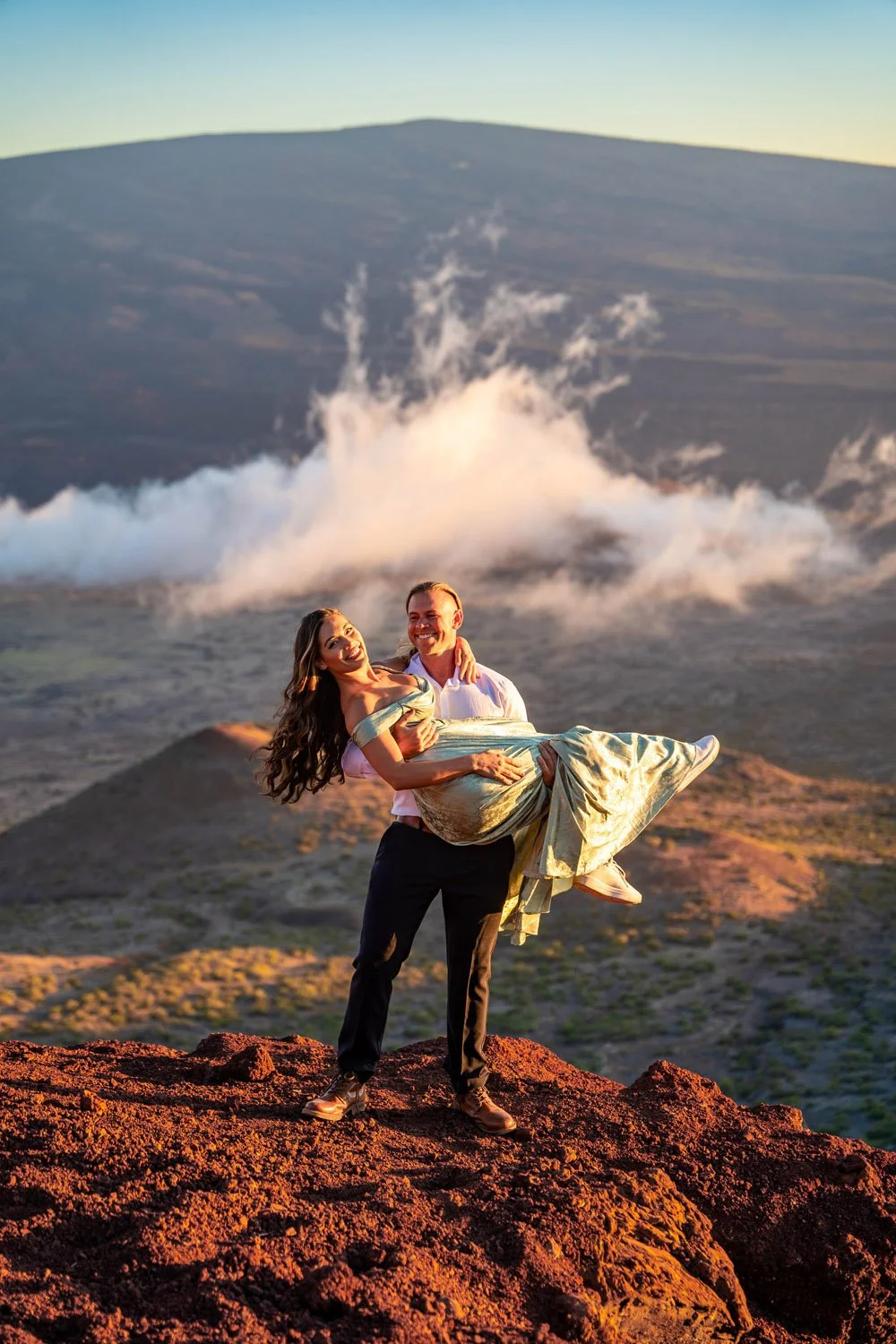 man carrying his girlfriend on a ledge with a mountain backdrop during sunset for a couples portrait session on the Big Island of Hawaii
