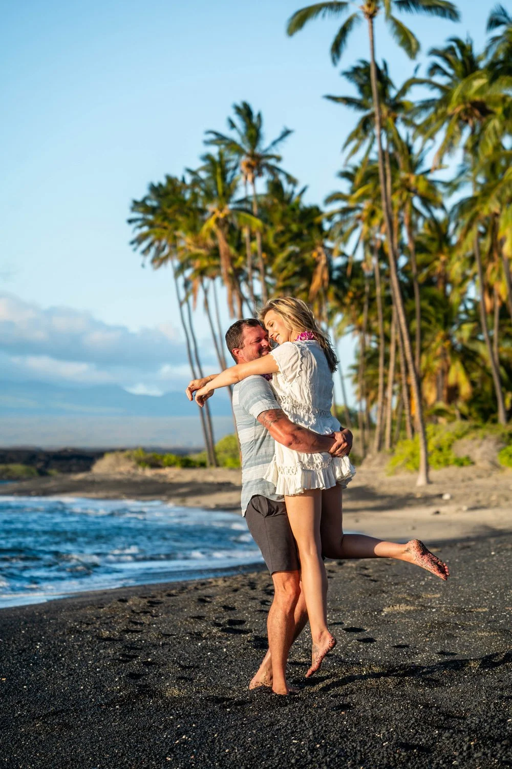 man holding his fiance girlfriend on a black sandy beach with palm trees in the background for an engagement couples portrait session on the Big Island of Hawaii
