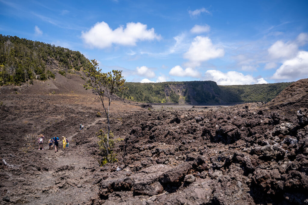 Visitors hiking across the hardened lava floor of Kīlauea Iki Crater under a bright blue sky in Hawaii Volcanoes National Park.
