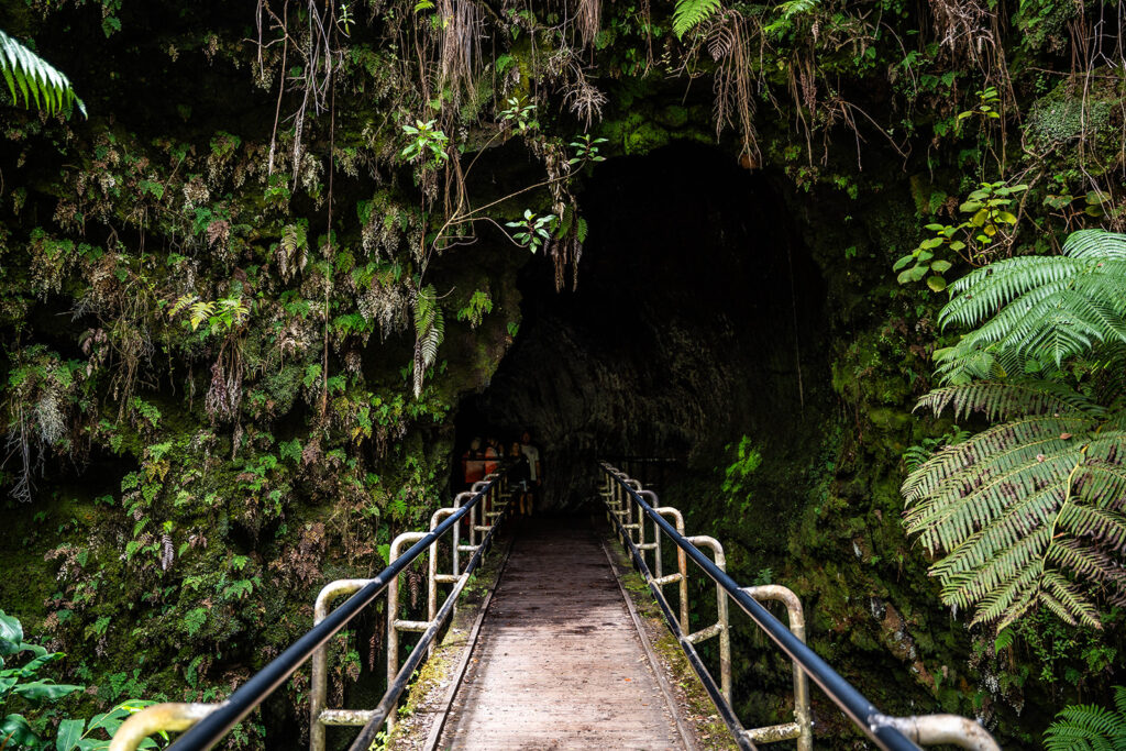 A narrow footbridge with railings leads into a dark, moss-covered cave entrance surrounded by lush green plants and ferns—a must-see stop on any Hawaii volcano itinerary.