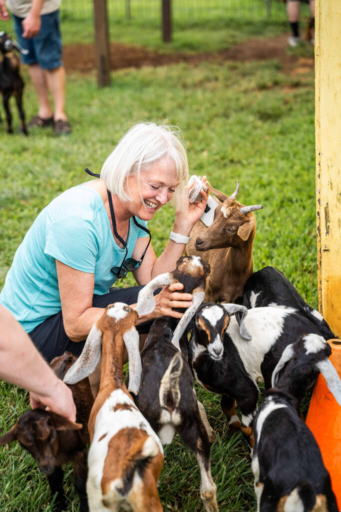 A smiling older woman kneels on grass, happily petting a group of friendly goats at the Honomu Goat Dairy Farm—one of the best easy half day trips for families with young kids on the Big Island. Other people and goats are visible in the background.