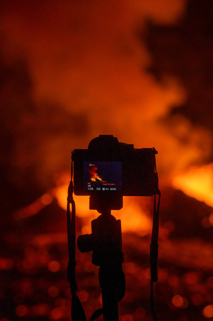A camera on a tripod is silhouetted against intense orange flames, its display capturing the eruption—a must-see moment for any Hawaii volcano itinerary or list of Hawaii Volcanoes National Park things to do.