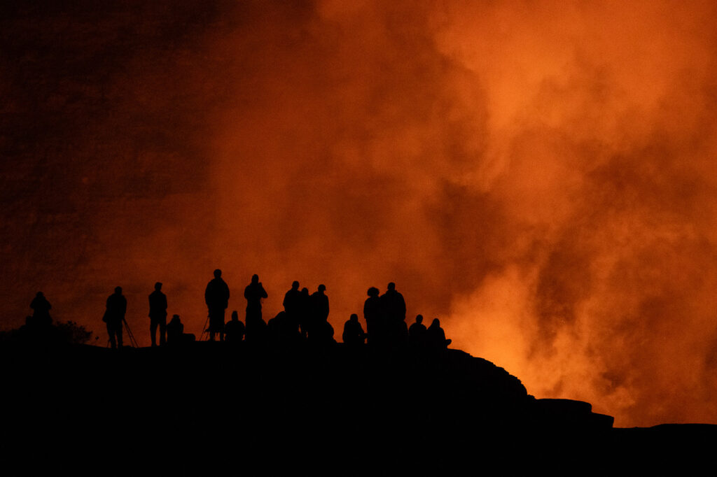 Silhouettes of visitors watching the glowing Halemaʻumaʻu Crater at night in Hawaii Volcanoes National Park.