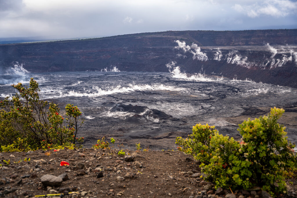 A steaming volcanic crater with dark, rocky terrain and sparse vegetation in the foreground, under a cloudy sky—an iconic scene and a must-see on any Hawaii volcano itinerary.