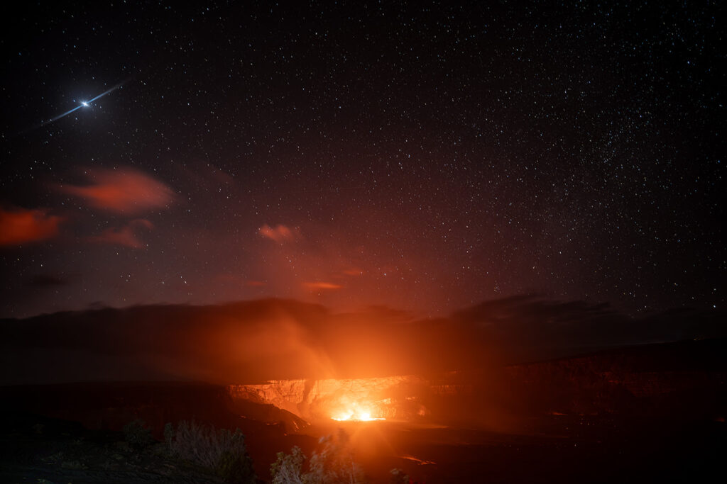 A glowing volcano emits bright orange light and smoke under a star-filled night sky, with a crescent moon shining above.