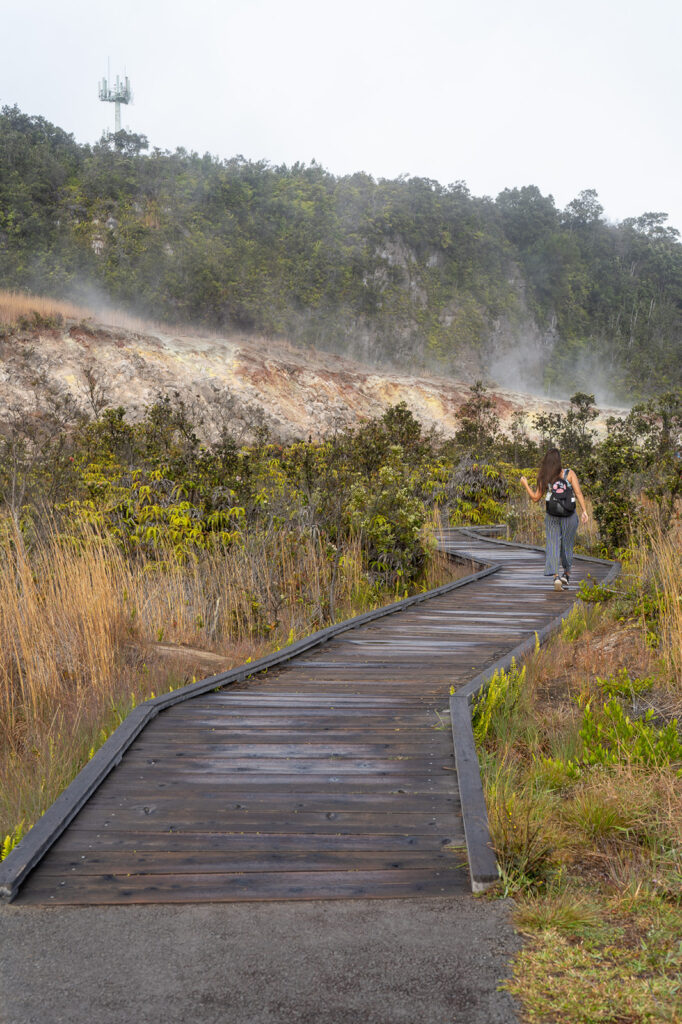Woman walking along the Sulfur Banks Trail boardwalk surrounded by steam and colorful volcanic cliffs in Hawaii Volcanoes National Park.