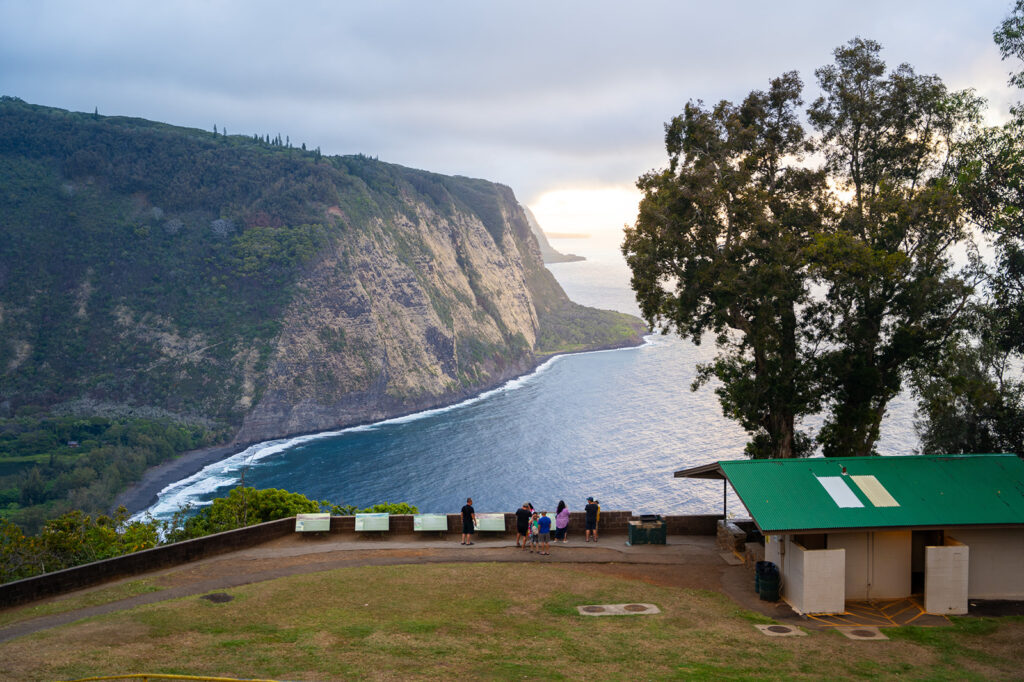 People stand at the Waipio Valley Lookout, enjoying a lush green cliff and coastline. Ocean waves curve along the shore, with a small green-roofed building in front—a perfect spot for easy half day trips on the Big Island.