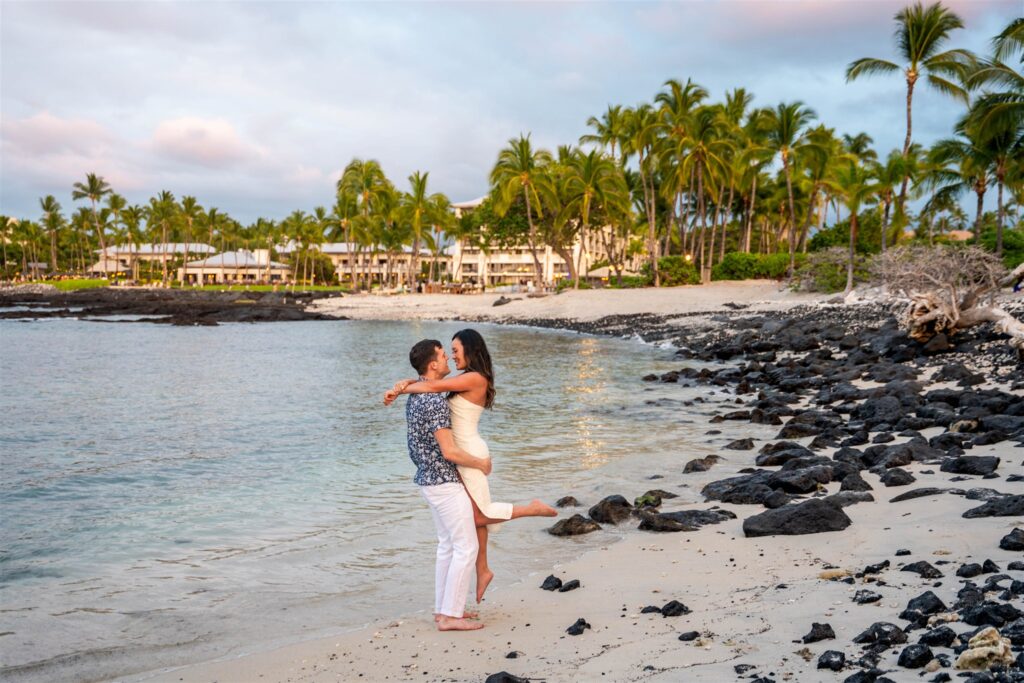 Couple embracing on the shoreline with palm trees and resort view after their sunset proposal at the Fairmont Orchid in Waikoloa, Hawaii.