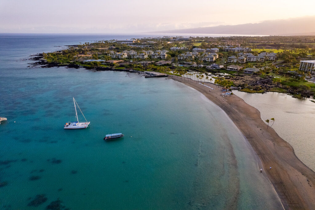 Aerial view of a coastal resort with turquoise water, sandy beach, and two boats anchored near shore; the lush green landscape makes it a dreamy spot for an Anaehoomalu Beach proposal under a mostly clear sky.
