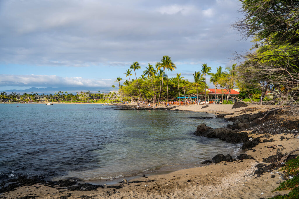 A sandy beach with dark rocks, palm trees, and clear blue water. People are relaxing near a beachfront restaurant, with resort buildings and mountains visible&mdash;an ideal spot for an Anaehoomalu Beach proposal under a partly cloudy sky.