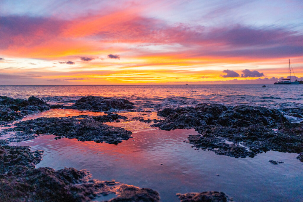A vibrant sunset over the ocean with colorful clouds reflected in the water and rocky shoreline in the foreground, capturing the romantic ambiance of an Anaehoomalu Beach proposal. A sailboat is visible near the horizon.