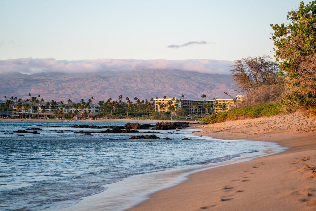 Golden sand beach with gentle waves and footprints leading along the shore, bordered by green shrubs&mdash;the perfect setting for an Anaehoomalu Beach proposal. In the distance are hotels, palm trees, and a mountain partially veiled by clouds under a blue sky.