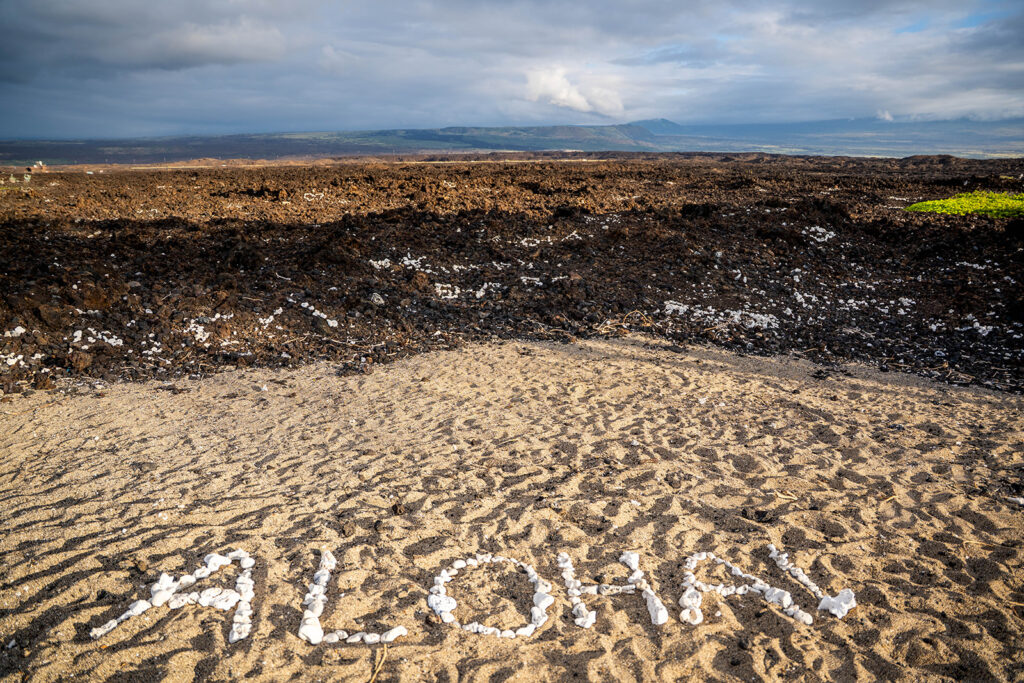 White rocks arranged to spell ALOHA on a sandy beach at Anaehoomalu, with dark volcanic rocks and mountains in the background&mdash;an idyllic spot for an Anaehoomalu Beach proposal under a partly cloudy sky.