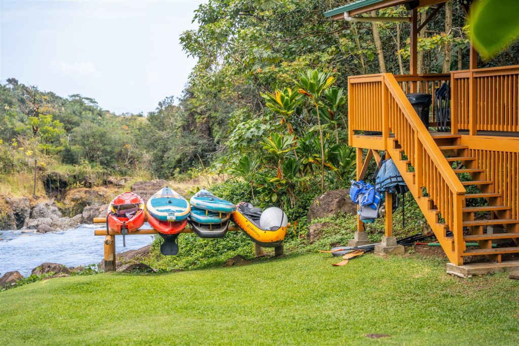 Colorful kayaks are stored on a wooden rack beside a cabin with stairs—an inviting setting for a hawaii horseback proposal. Lush green grass, tropical plants, and a rocky river complete the scene. Life jackets and paddles hang near the cabin.