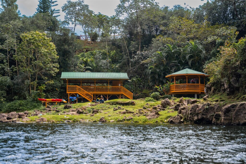 A riverside scene shows two yellow wooden structures with green roofs surrounded by lush trees. Kayaks rest on the grass near rocky banks, and people gather under the gazebos beside the water, creating a setting perfect for a Hawaii horseback proposal.