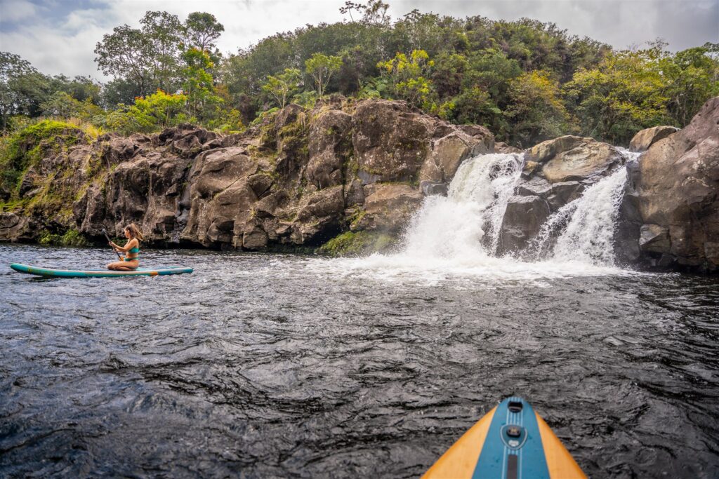 A person on a paddleboard faces a small waterfall surrounded by rocky cliffs and lush greenery, hinting at adventure and romance—reminiscent of a Hawaii horseback proposal—with the tip of another paddleboard visible in the foreground.