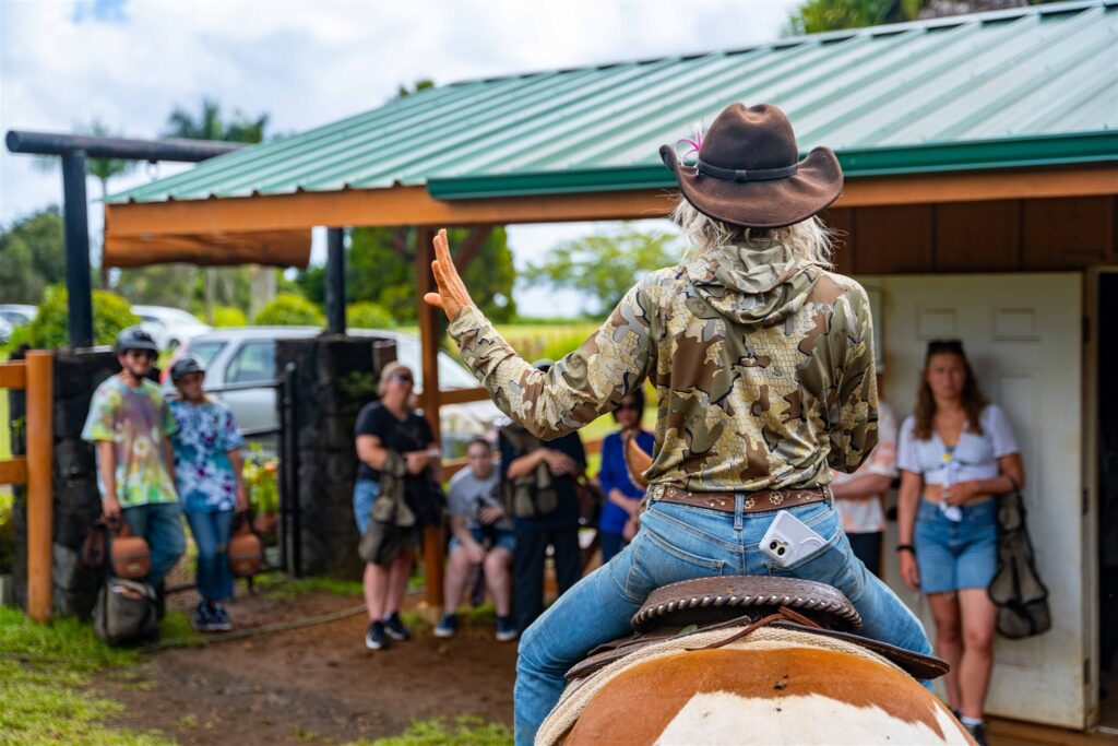 A person in a cowboy hat and camouflage shirt sits on a horse, gesturing with their hand while speaking to a group of people outside a green-roofed building, setting the perfect scene for a unique Hawaii horseback proposal.