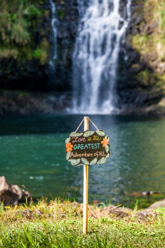 A small sign saying Love is the GREATEST Adventure of ALL stands on grass near a calm, blue-green pond with a waterfall in the background&mdash;a dreamy setting perfect for unique Hawaii proposal ideas.