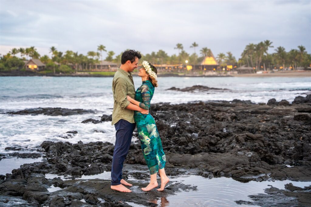 A couple stands barefoot on black volcanic rocks by the ocean, embracing and touching noses&mdash;a perfect scene for hawaii proposal ideas. The woman wears a green dress and flower crown; the man wears a green shirt. Palm trees and huts line the shore behind them.