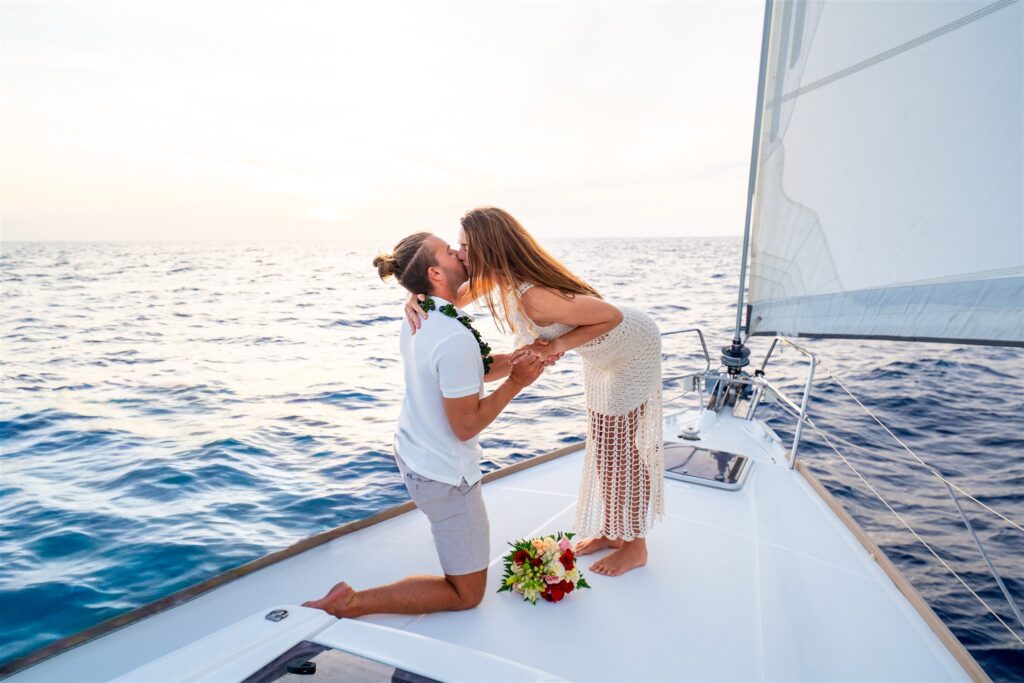 A couple on a sailboat at sunset, with the man kneeling and holding the woman's hands as they kiss. Both wear leis, surrounded by flower bouquets and calm ocean water&mdash;one of the most romantic Hawaii proposal ideas.