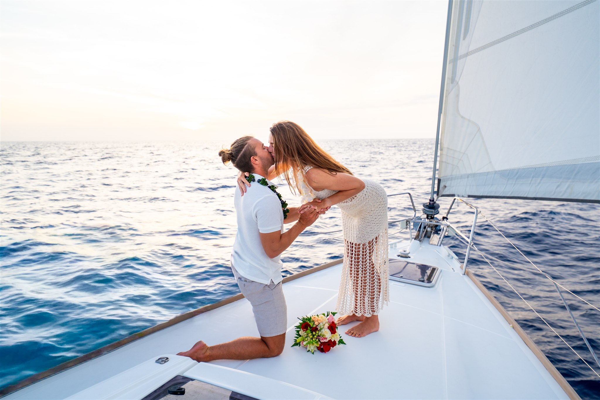 A couple on a sailboat at sunset, with the man kneeling and holding the woman's hands as they kiss. Both wear leis, surrounded by flower bouquets and calm ocean water—one of the most romantic Hawaii proposal ideas.