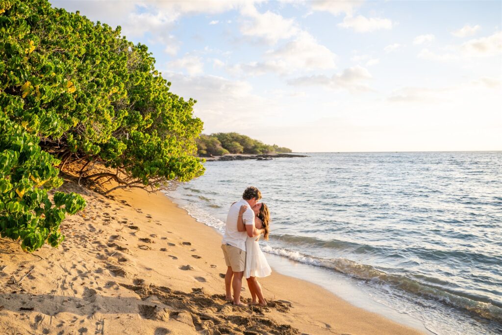 A couple stands on a sandy beach at sunset, embracing and kissing near the shore after an Anaehoomalu Beach proposal. Green trees line the left side, gentle waves wash up, and footprints are left in the sand beneath a partly cloudy sky.