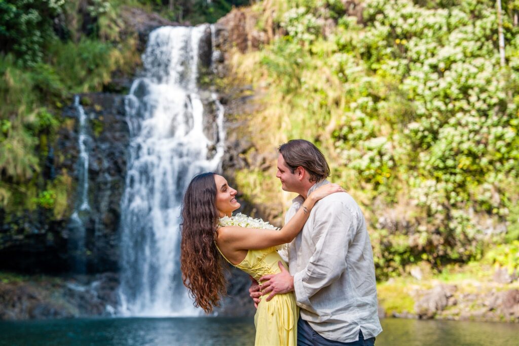 A couple embraces and smiles at each other in front of a lush waterfall, surrounded by greenery&mdash;an idyllic scene perfect for hawaii proposal ideas. The woman wears a yellow dress and flower lei; the man wears a light-colored shirt.