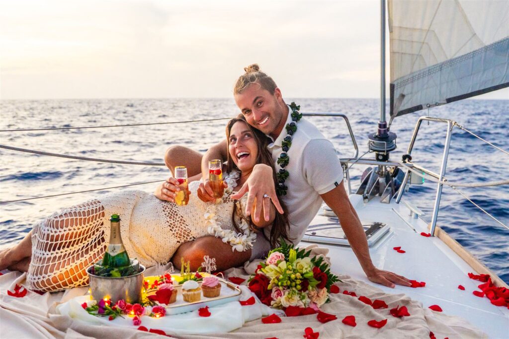 A joyful couple celebrates on a sailboat at sea, surrounded by rose petals, champagne, and desserts&mdash;one of the sweetest Hawaii proposal ideas&mdash;as the woman shows off her engagement ring and both smile brightly.