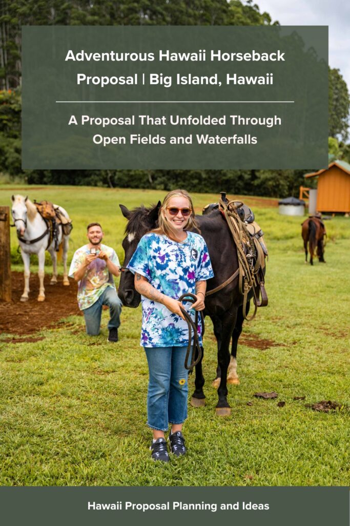 A smiling woman stands beside a saddled horse, holding its reins during a romantic Hawaii horseback proposal, while a man kneels behind her on the grass with a ring box. Other horses and a small building are visible in the background.