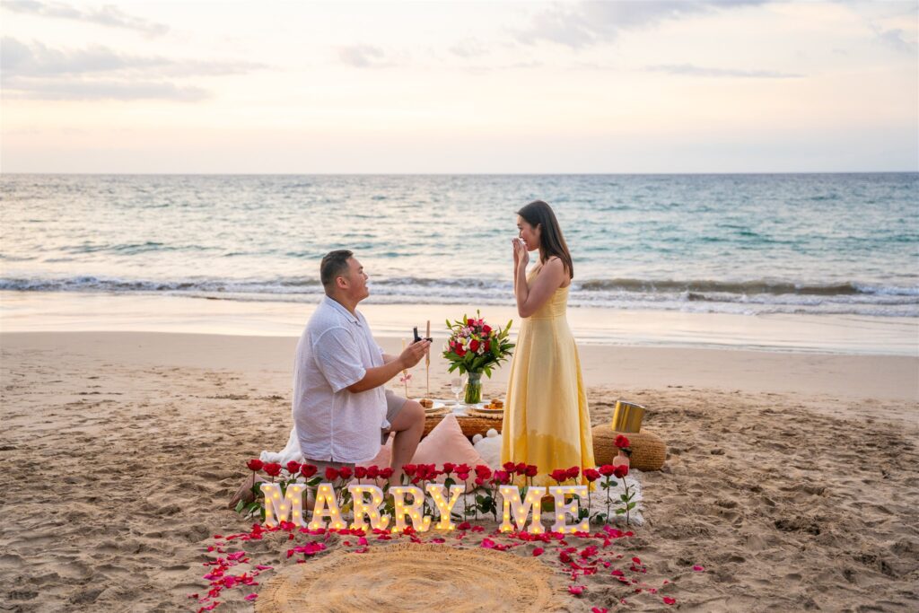 A man kneels on a beach holding a ring box, proposing to a surprised woman in a yellow dress. "MARRY ME" is spelled out in lights and red roses, with a sunset and ocean&mdash;perfect for those seeking Hawaii proposal ideas.