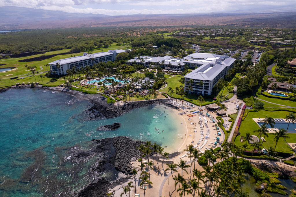 Aerial view of a large seaside resort, ideal for a Fairmont Orchid proposal, with several buildings, swimming pools, palm trees, and a sandy beach bordered by clear turquoise water. People are swimming and sunbathing amid lush greenery and distant hills.