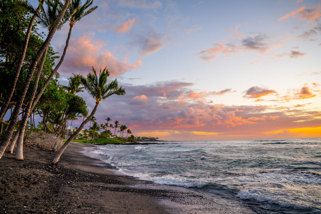 Palm trees line a sandy beach as gentle waves meet the shore under a colorful sunset sky, creating a romantic setting perfect for a Fairmont Orchid proposal. The ocean stretches into the distance, with lush greenery bordering the coastline.