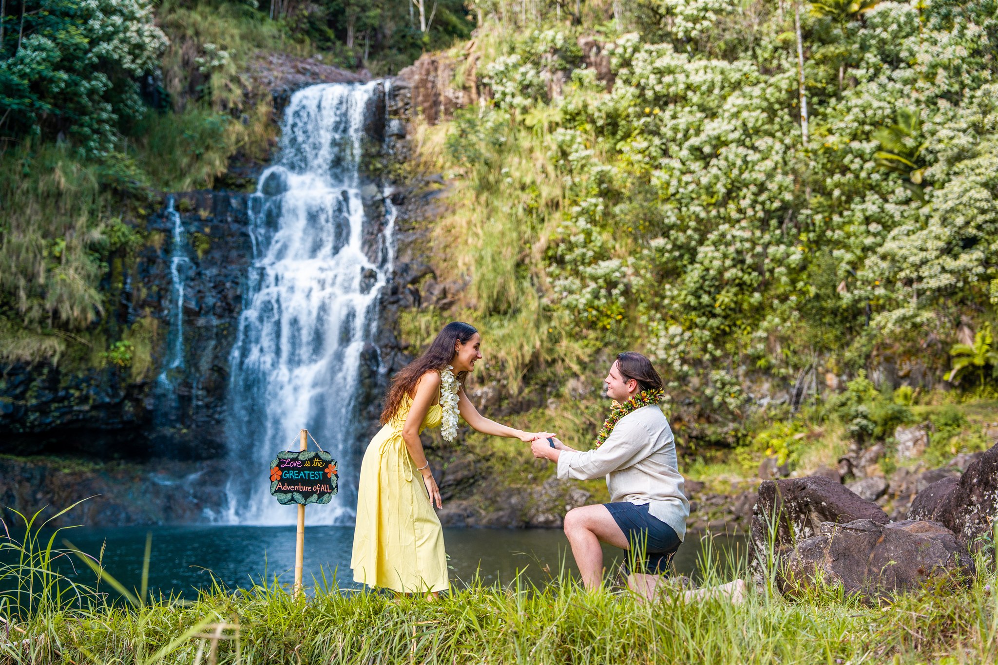 A man kneels and holds a woman's hand in front of a waterfall surrounded by lush greenery&mdash;a dreamy scene for those seeking Hawaii proposal ideas. The woman wears a yellow dress; the man is in a light shirt and shorts. A small colorful sign stands on the grass nearby.