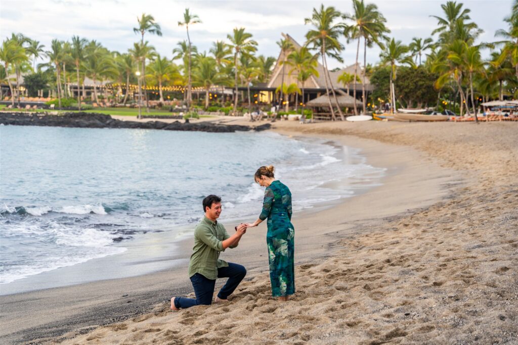 A man kneels on a sandy beach, proposing to a woman standing before him&mdash;a romantic scene perfect for those seeking unique Hawaii proposal ideas. Palm trees and buildings line the shoreline in the background as she admires the ring on her hand.