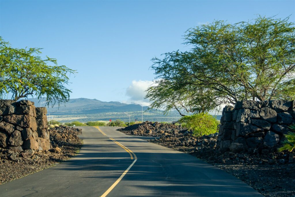 A paved road curves through a rocky landscape with stone walls and green trees on both sides near Kikaua Point Park, leading toward distant mountains under a clear blue sky.