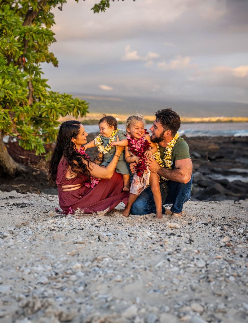 Family crouches down together on the sand, smiling at each other during Hawaiian family photos.