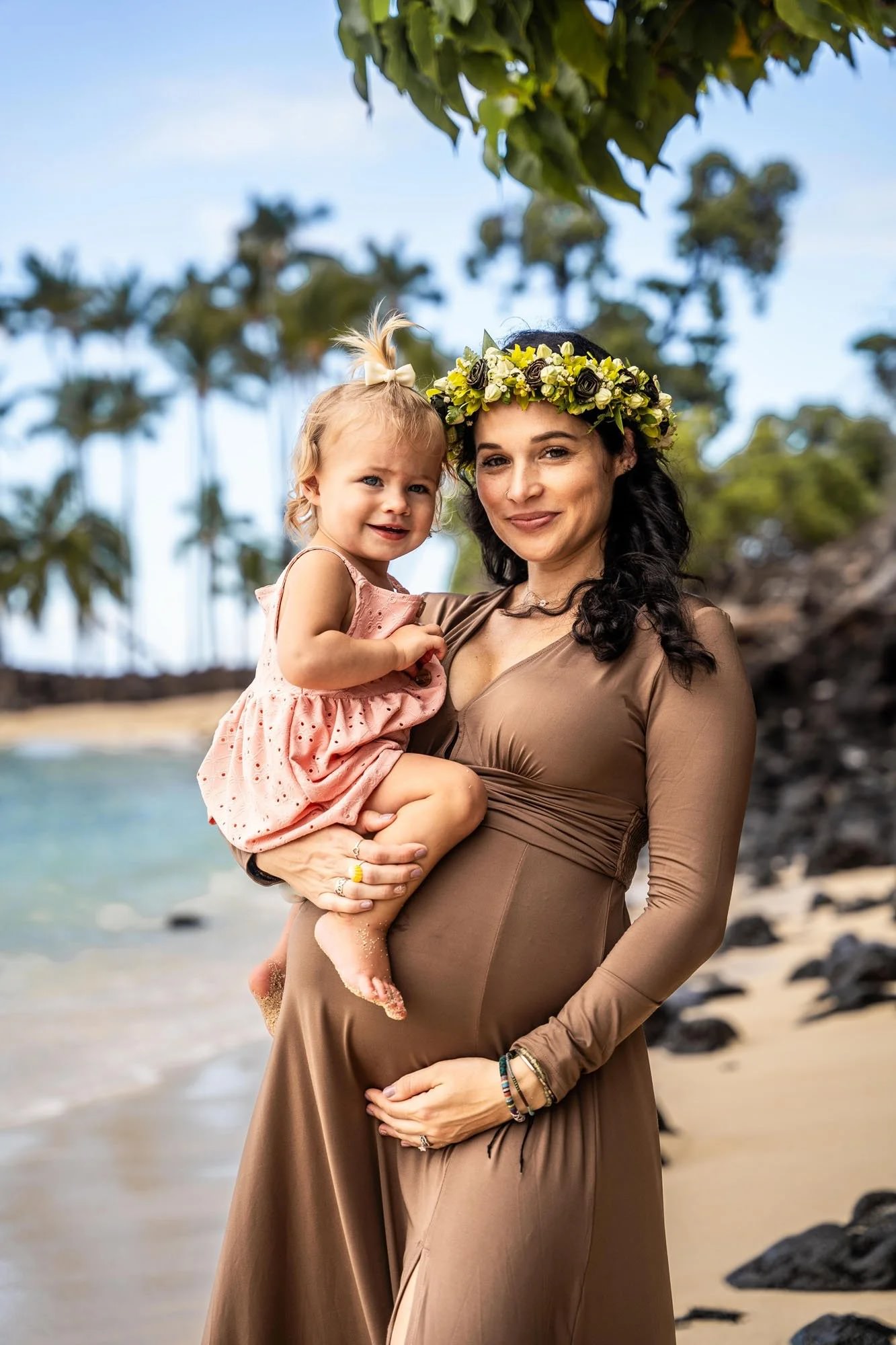 Pregnant mother holding her toddler during a Hawaiian family photoshoot on a tropical beach.